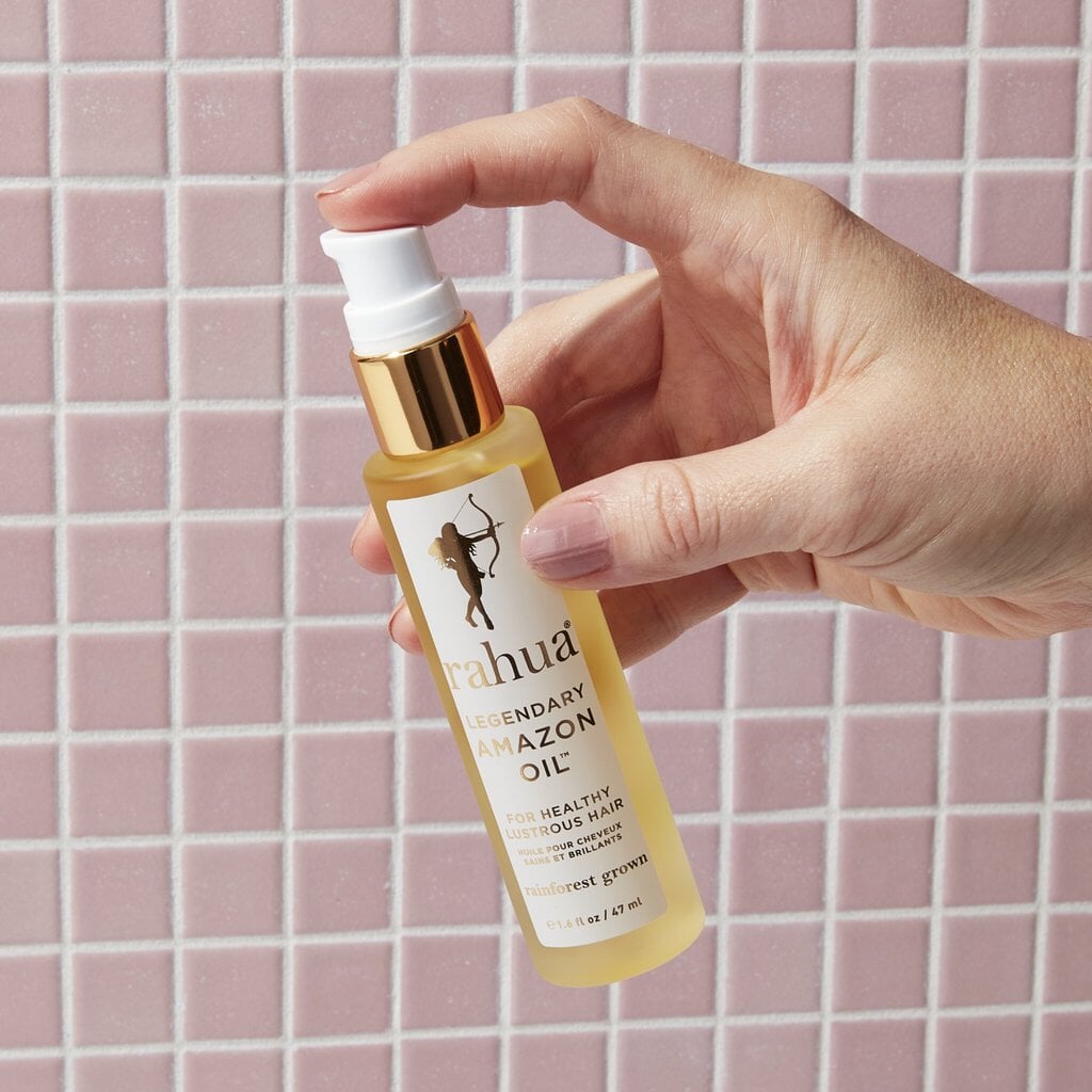 Rahua Legendary Amazon Oil Flasche gehalten von einer Hand vor rosafarbenen Fliesenhintergrund.