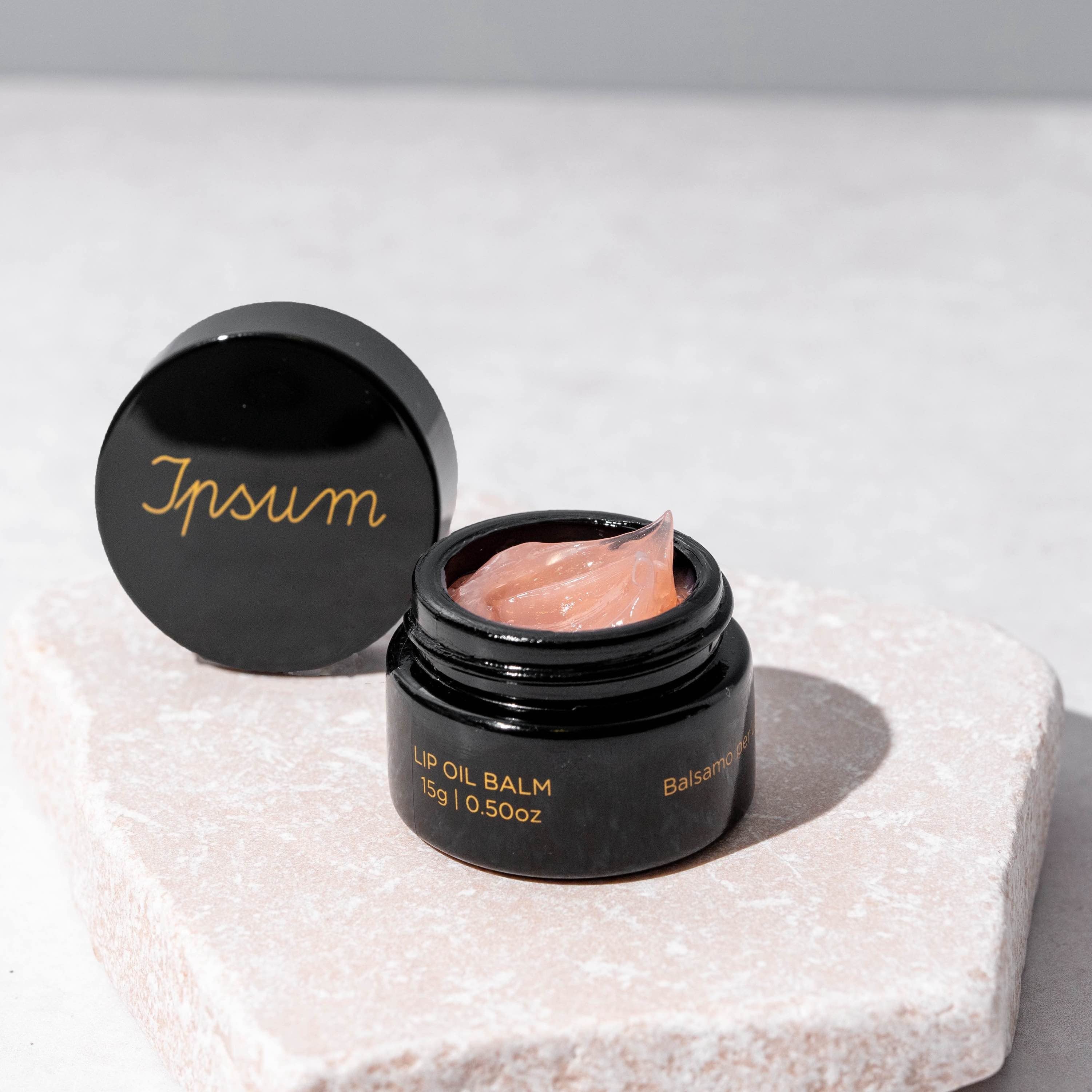 Schwarzer Ipsum Lip Oil Balm Tiegel steht offen mit angelehntem Deckel auf einer Steinplatte, Balm im zarten Rosa und gescheidiger Textur gut zu erkennen.
