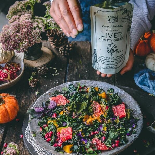 Anima Mundi Liver Verpackung gehalten von einer Hand, die andere Hand streut grünes Pulver auf eine Teller mit buntem Gemüse.