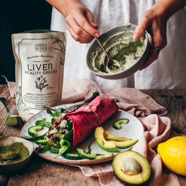 Anima Mundi Liver Verpackung steht geöffnet auf einem Tisch bei einem Teller auf dem ein Wrap mit viel Gemüse liegt, im Hintergrund ein Ausschnitt eines Menschen, der einen Dip in einer Schale mit dem Liver Pulver anrührt.
