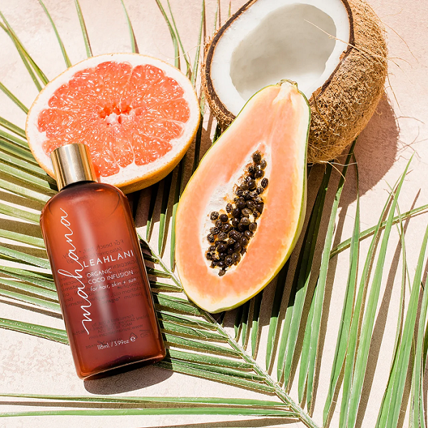 Leahlani Mahana Körperöl Flasche auf grünem Palmenblatt mit aufgeschnittener Orange, Papaya und Kokosnuss