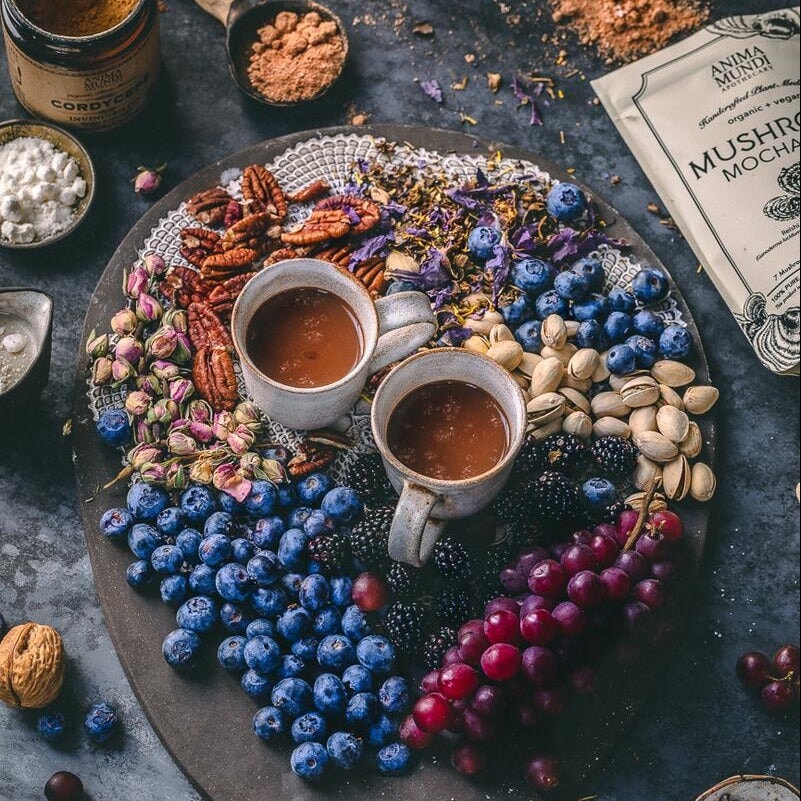 2 Tassen mit Mushroom Mocha Milk stehen in der Mitte umringt von Baubeeren, Nüssen, und getrockneten Blüten, eine Anima Mundi Mushroom Moha Milk Tüte liegt dahinter. North Glow