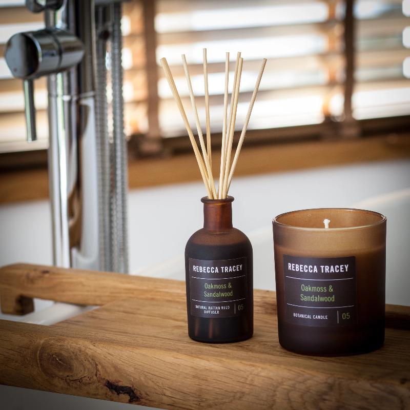 Stäbchen Diffusor und Kerze "Oakmoss & Sandalwood" nebeneinander stehend auf einer Badewannenablage aus Holz.