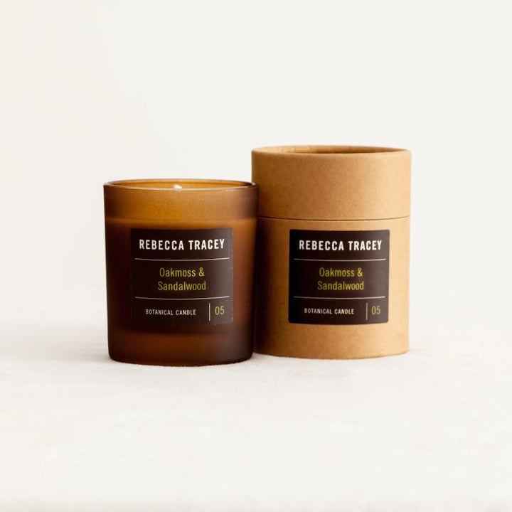 Kerzenglas und Verpackung "Oakmoss & Sandalwood" von Rebecca Tracey stehend nebeninander vor weißem Hintergrund.