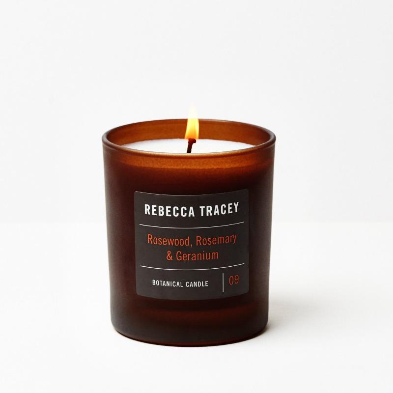 Brennende Kerze von Rebecca Tracey in braunem Glas in den Duftnoten Rosewood, Rosemary & Geranium vor weißem Hintergrund.