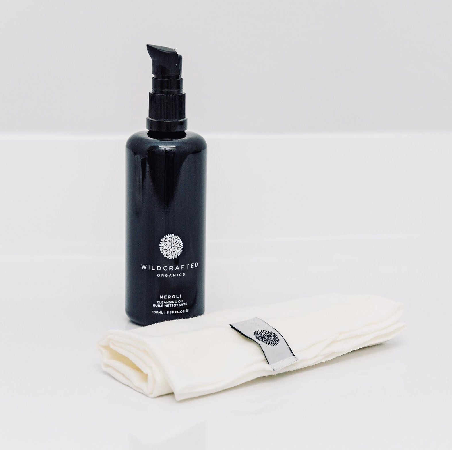Schwarze Pumpflasche von Wildcrafted Organics Neroli Cleanser vor weißem Hintergrund, davor liegt eine Musselinreinigungstuch.