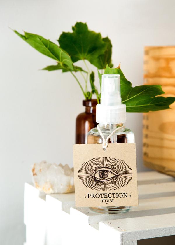 Anima Mundi Palo Santo Glasflasche mit einem großen Papierschild auf dem ein großes Auge abgebildet ist steht auf einem Regal, im Hintergrund grüne Blätter in einer Vase.