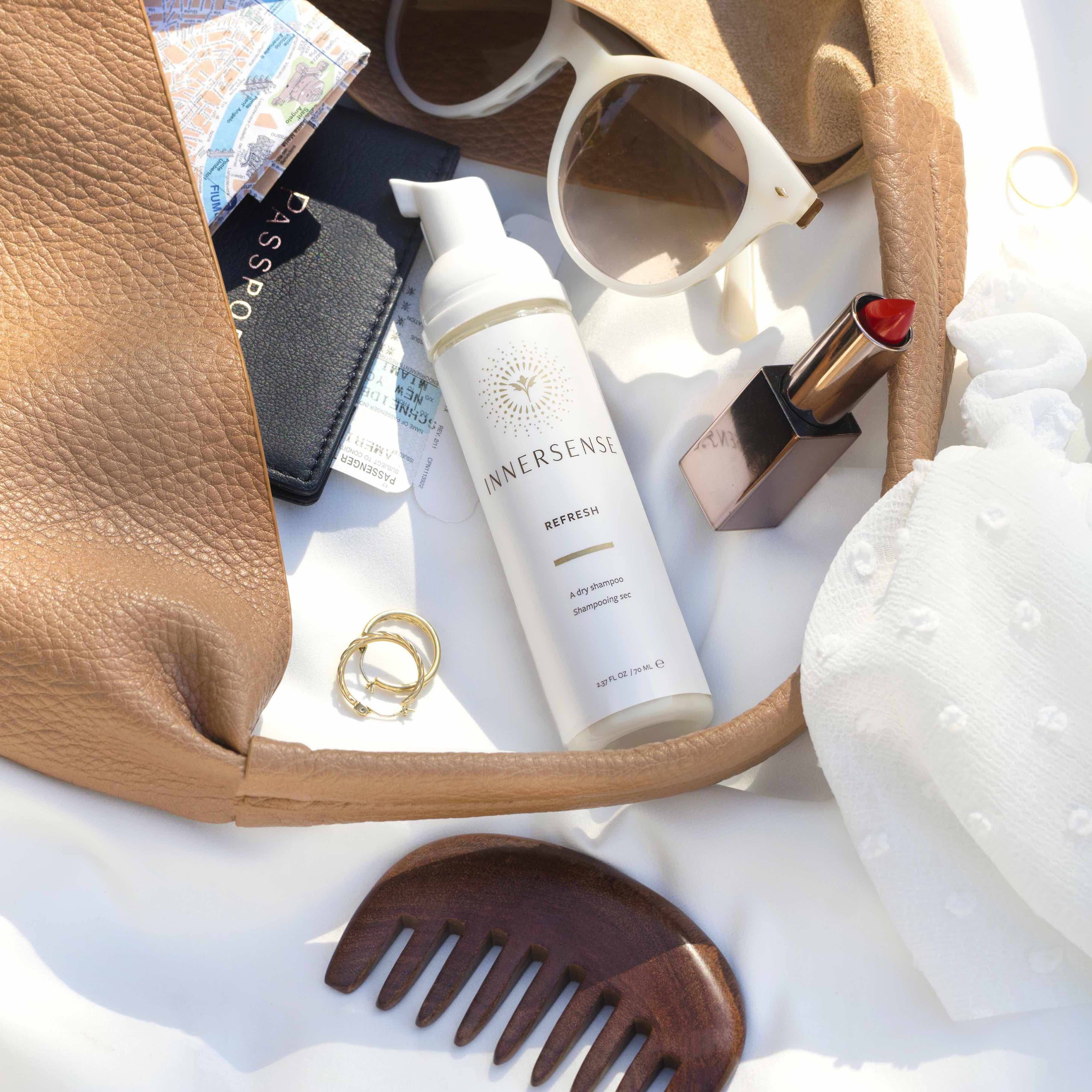 Pumpflasche Innersense Refresh Flasche liegt vor einer ausgekippten Handtasche zwischen Sonnenbrille, Lippenstift, Ohrringen und Holzkamm.