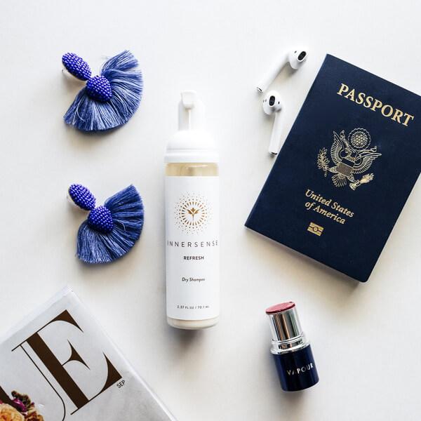 Pumpflasche Innersense Refresh Flasche liegt zwischen einem Reisepass, Lippenstift, Airpods und blauen Ohrringen.