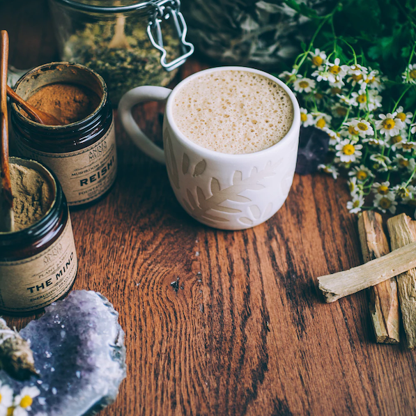 Anima Mundi Schraubglas Reishi steht neben einer Tasse mit cremiger Flüssigkeit auf einem Holzuntergrund zwischen Kräutern und Palo Santo Holz.