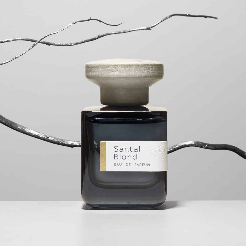 Dunkelblaue Glasflasche mit grauem Deckel  "Santal Blond" von Atelier Materi mit einem silbernen Ast im Hintergrund.
