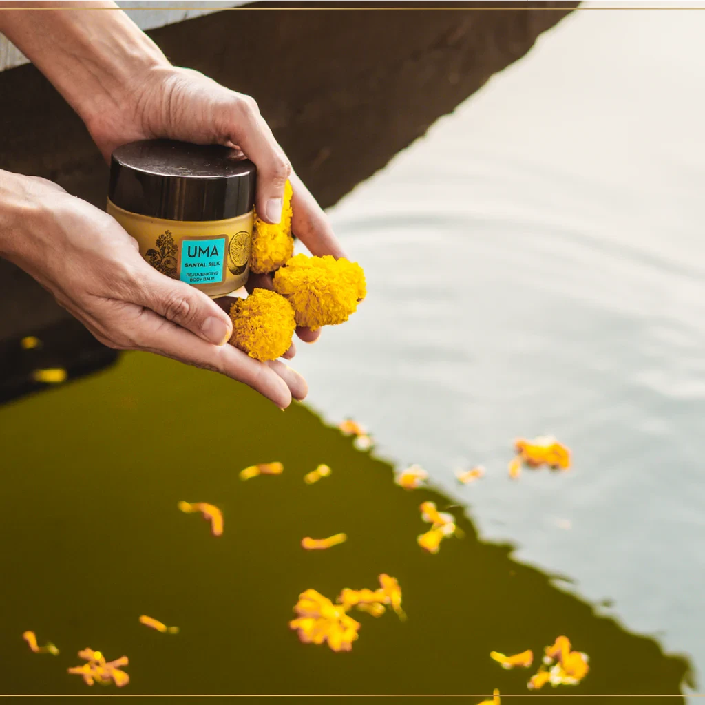 2 Hände halten Blütenblätter und ein Schraubglas des UMA Santal Body Balms über reflektierendem Wasser in dem Blütenblätter schwimmen.