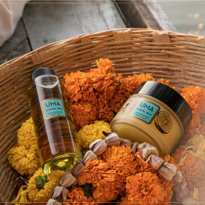 In einem Weidenkörbchen gefüllt mit orangefarbenen Blütenblättern liegen eine Flasche Santal Silk Body Oil und ein Schraubglas Santal Silk Body Balm.