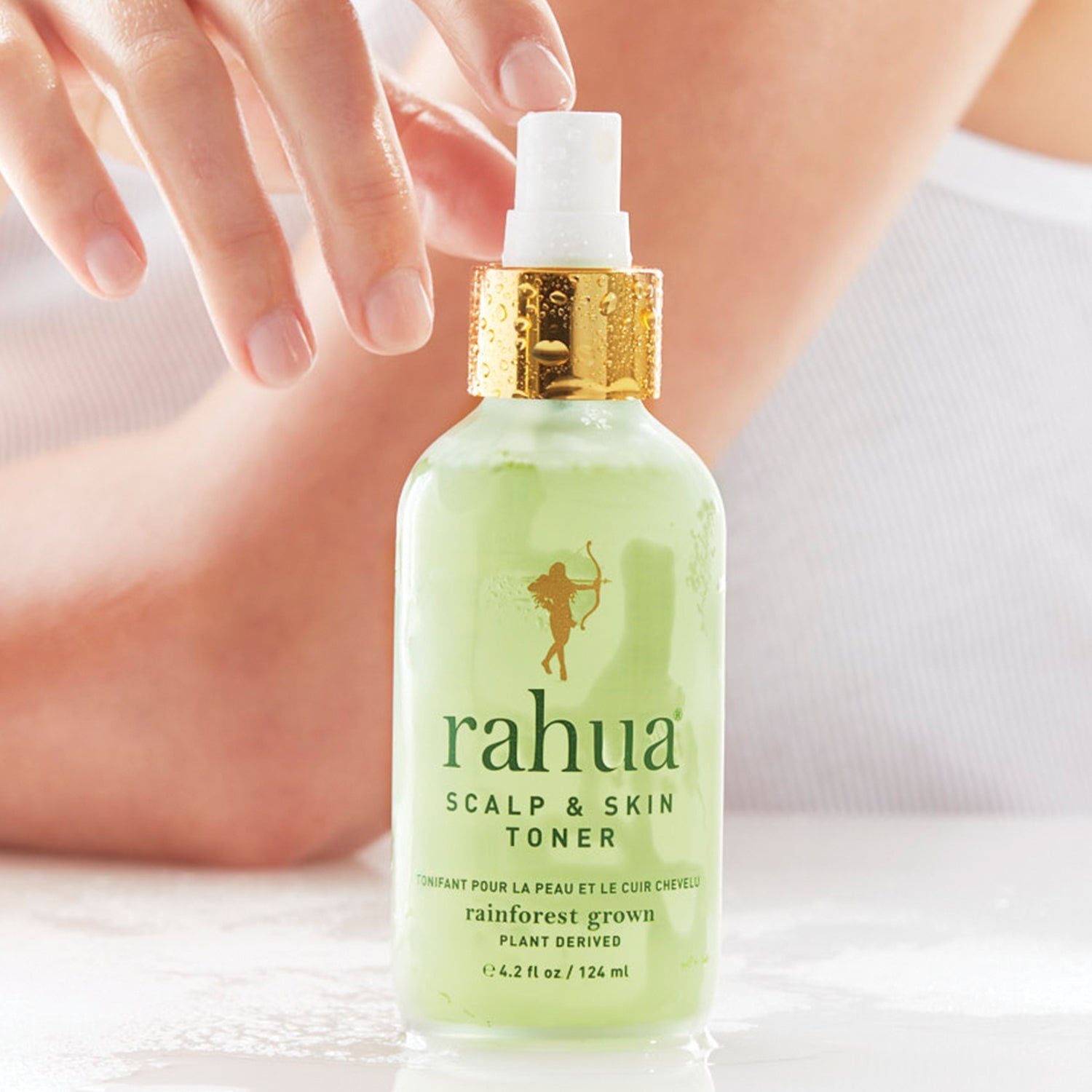Rahua Scalp & Skin Toner Flasche steht auf einem weißem Tisch, im Hintergrund verschwommen die Silhouette einer Frau.