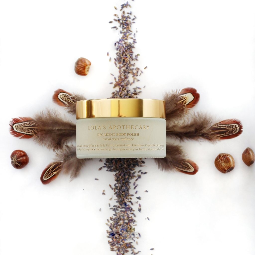 Ein Lola's Apothecary Body Polish Glastiegel mit goldenem Deckel liegt auf einem Bett aus Lavendelblüten und Federn.