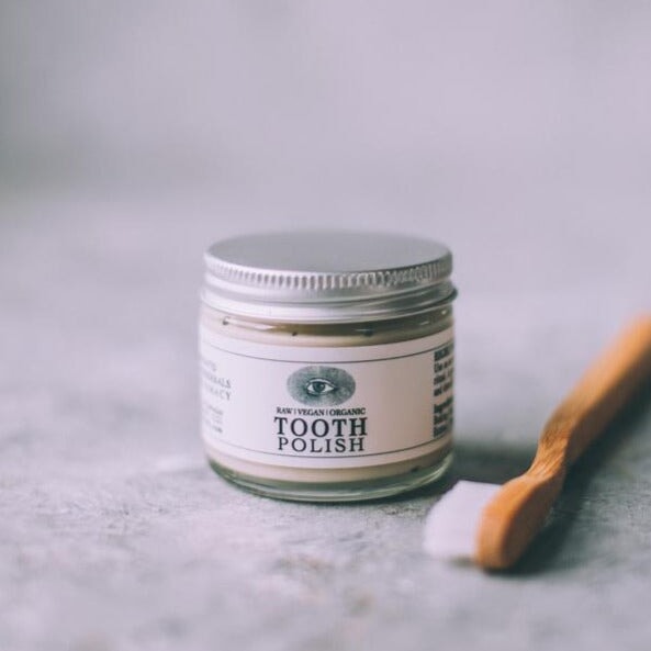 Schraubglas Toothpolish von Anima Mundi mit silbernen Deckel steht neben einer Zahnbürste aus Holz.