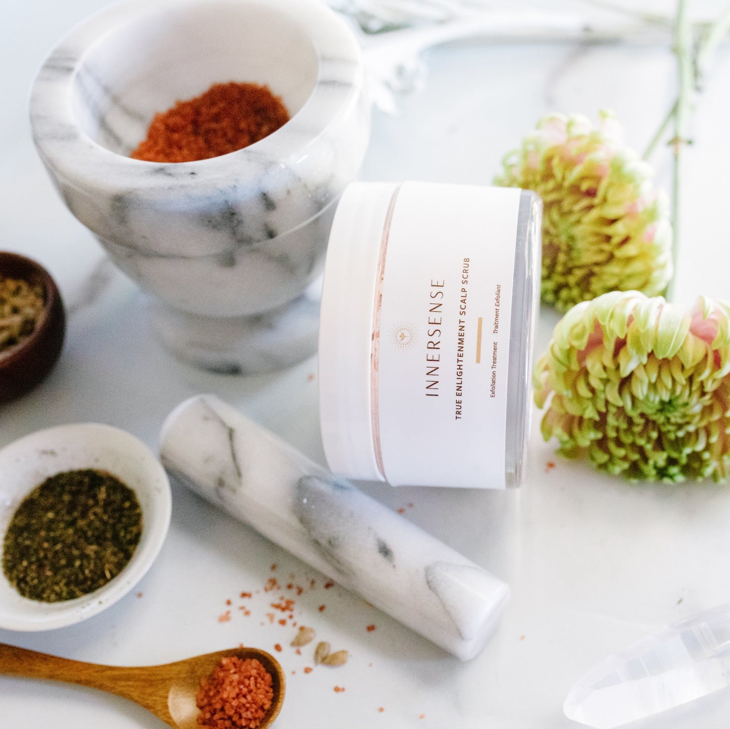 Innersense True Enlightenment Scalp Scrub Tiegel liegt seitlich neben einem Mörser und verschiedenen Kräutern in Schüsselchen zwischen Blüten. 