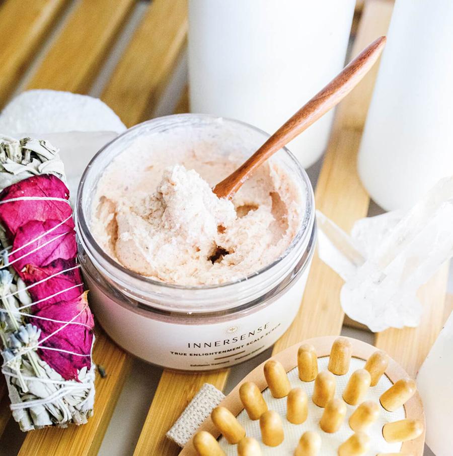 Offener Innersense True Enlightenment Scalp Scrub Tiegel mit einem Holzlöffel liegt neben einer Bürste mit dicken Borsten aus Holz auf einem Holzbrett