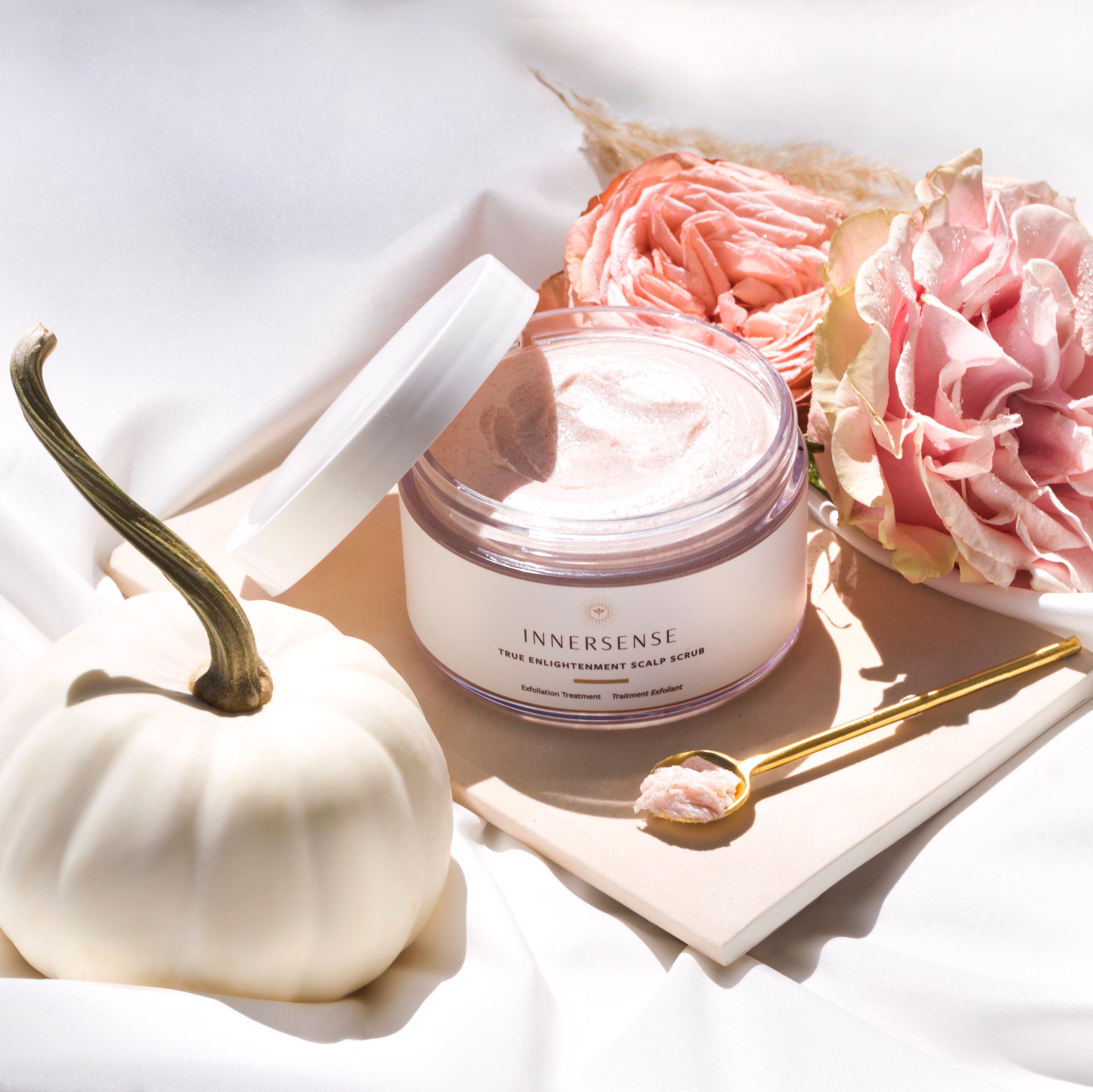 Innersense True Enlightenment Scalp Scrub Tiegel mit offenem Deckel dkoriert mit Messinglöffel und rosa Blüten auf hellem Hintergrund.