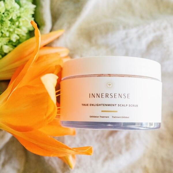 Innersense True Enlightenment Scalp Scrub Tiegel liegt neben einer orangefarbenen Blüte.