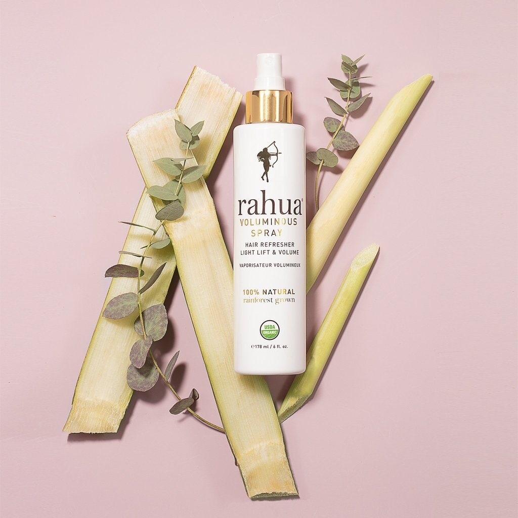 Rahua Voluminous Spray Flasche vor rosafarbenem  Hintergrund, dekoriert mit verschiedenen Blättern. North Glow