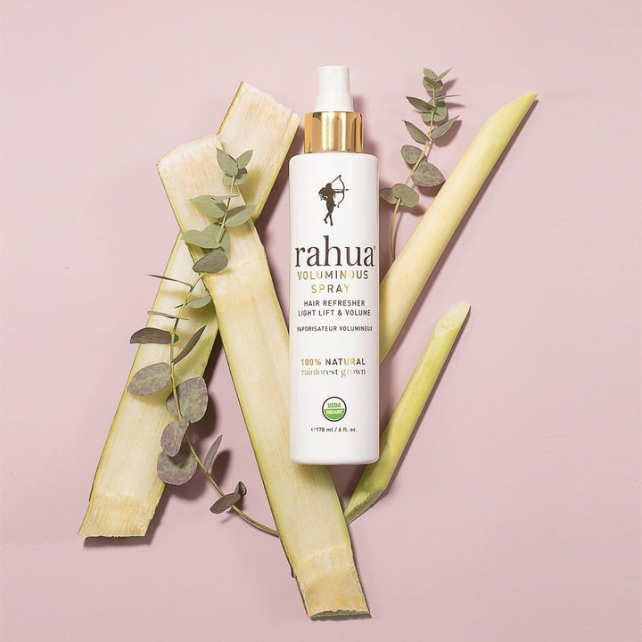 Rahua Voluminous Spray Flasche vor rosafarbenem  Hintergrund, dekoriert mit verschiedenen Blättern.