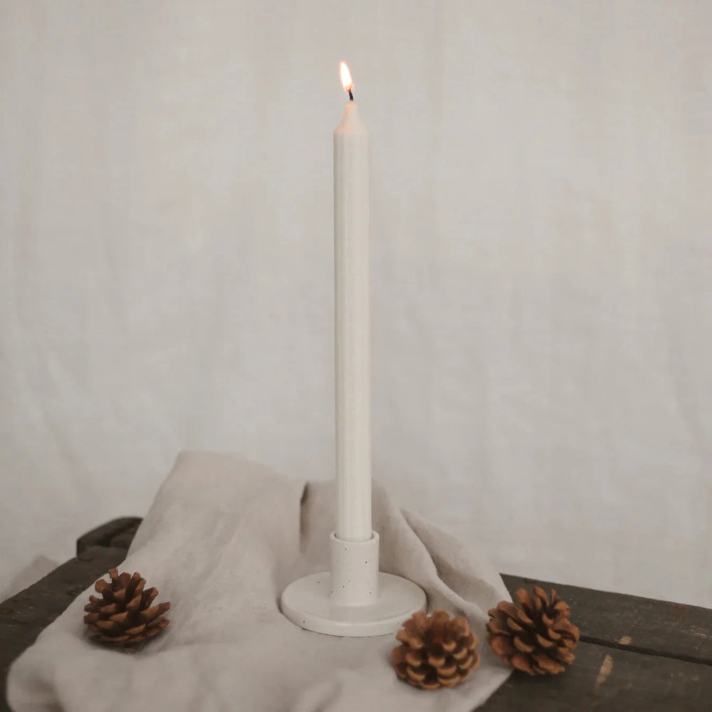 Lighted white stick candle by Eulenschnitt in a candle holder on a beige linen cloth, next to it 3 pine cones.