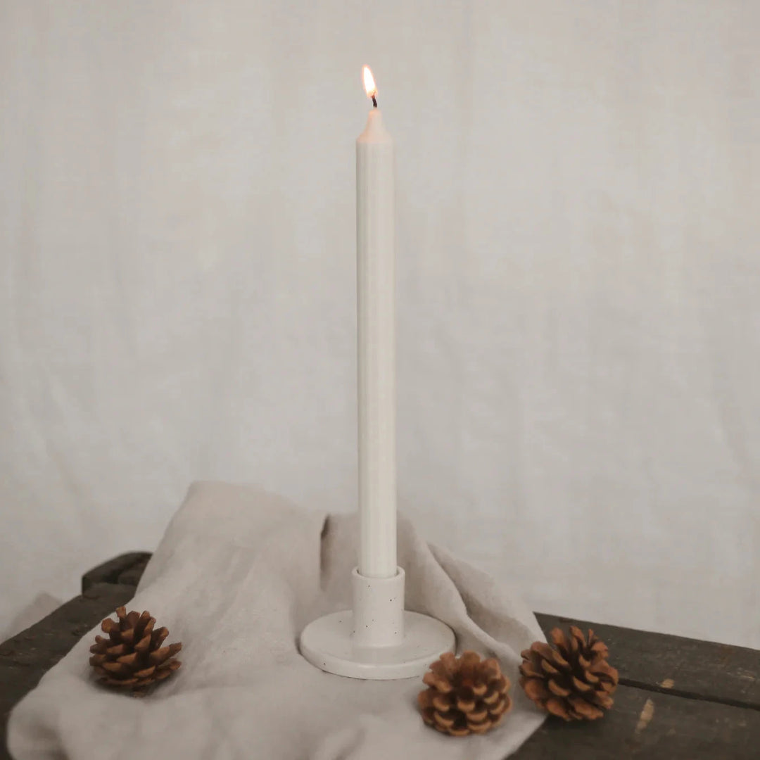 Lighted white stick candle by Eulenschnitt in a candle holder on a beige linen cloth, next to it 3 pine cones. North Glow