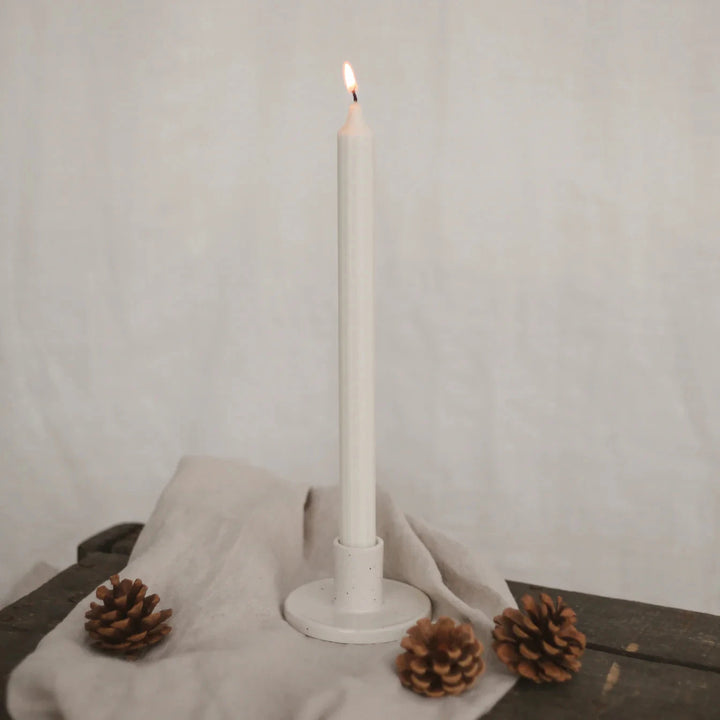 Lighted white stick candle by Eulenschnitt in a candle holder on a beige linen cloth, next to it 3 pine cones.