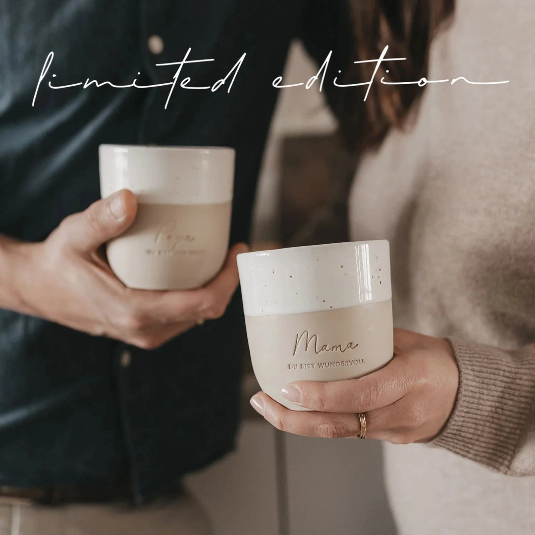 Becher "Mama du bist wundervoll" wird von einer Hand gehalten, dahinter leicht verschwommen wird ein anderer Becher in einer Hand gehalten. North Glow