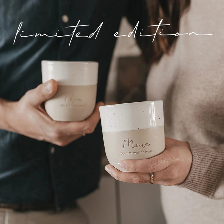 Becher "Mama du bist wundervoll" wird von einer Hand gehalten, dahinter leicht verschwommen wird ein anderer Becher in einer Hand gehalten.