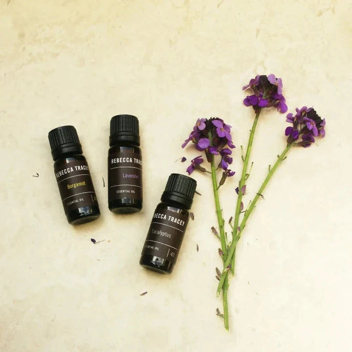 3 small dark brown glass bottles of Essential Oil by Rebeccy Tracey lying next to flowers.