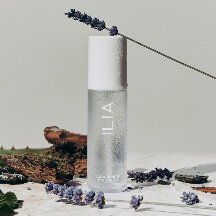 ILIA Blue Light Mist glass bottle with white lid is decorated with lavender on a light background.