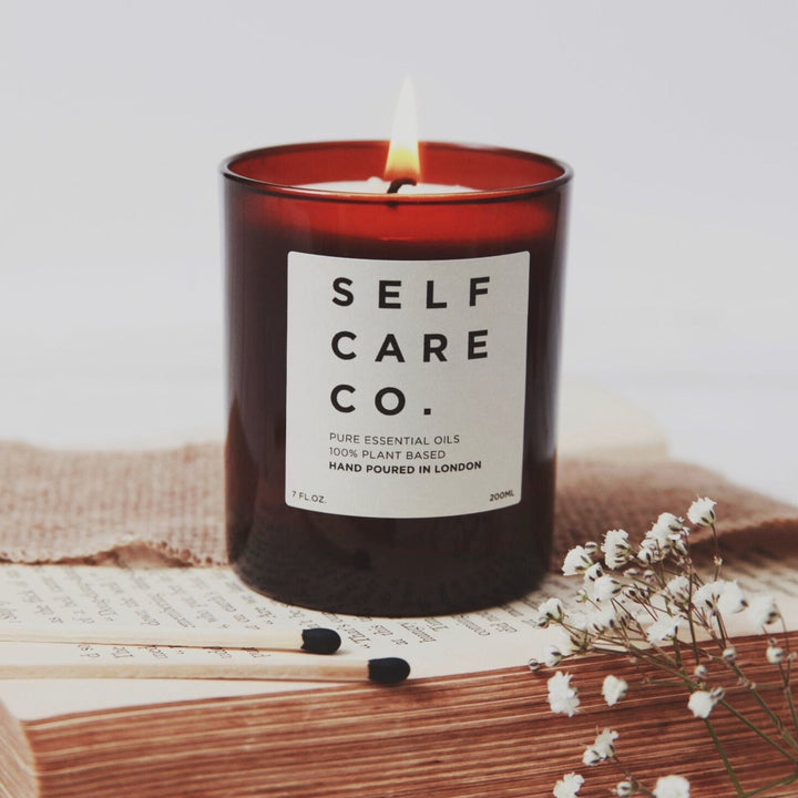 Brown candle jar with the inscription Self Care Co. stands on a book, in the foreground are two matches