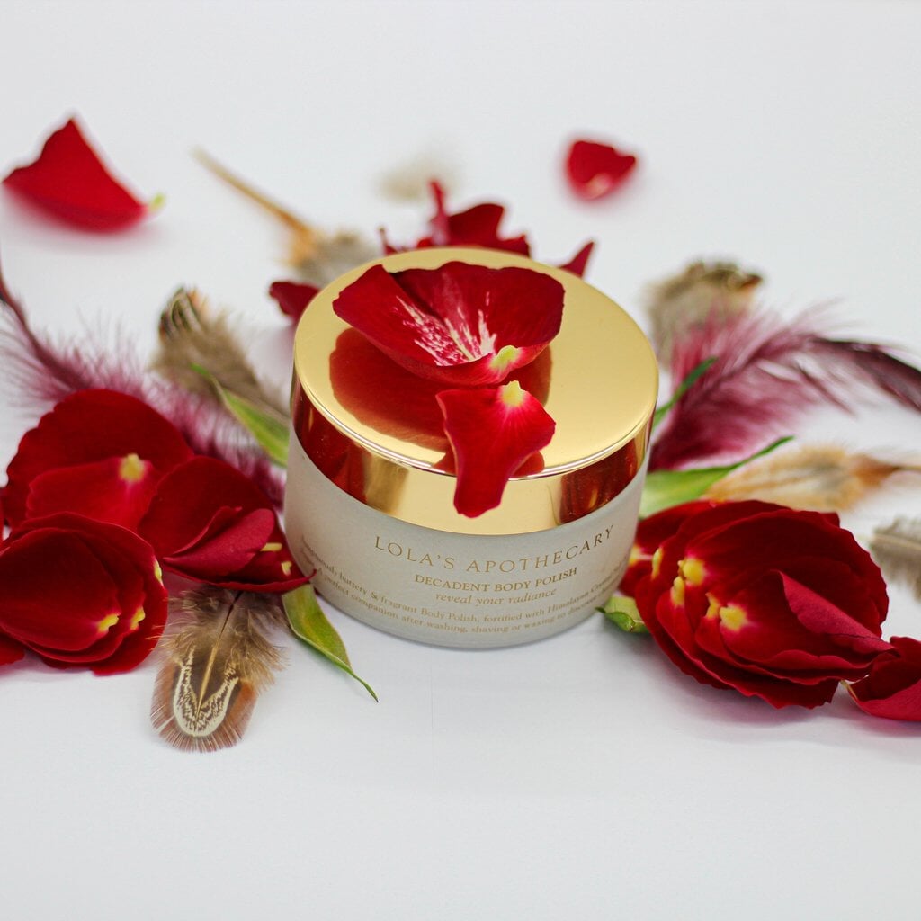 Glastiegel Lola's Apothecary Body Polish mit goldenem Deckel steht mit Rosenblättern und Federn dekoriert auf hellem Untergrund.