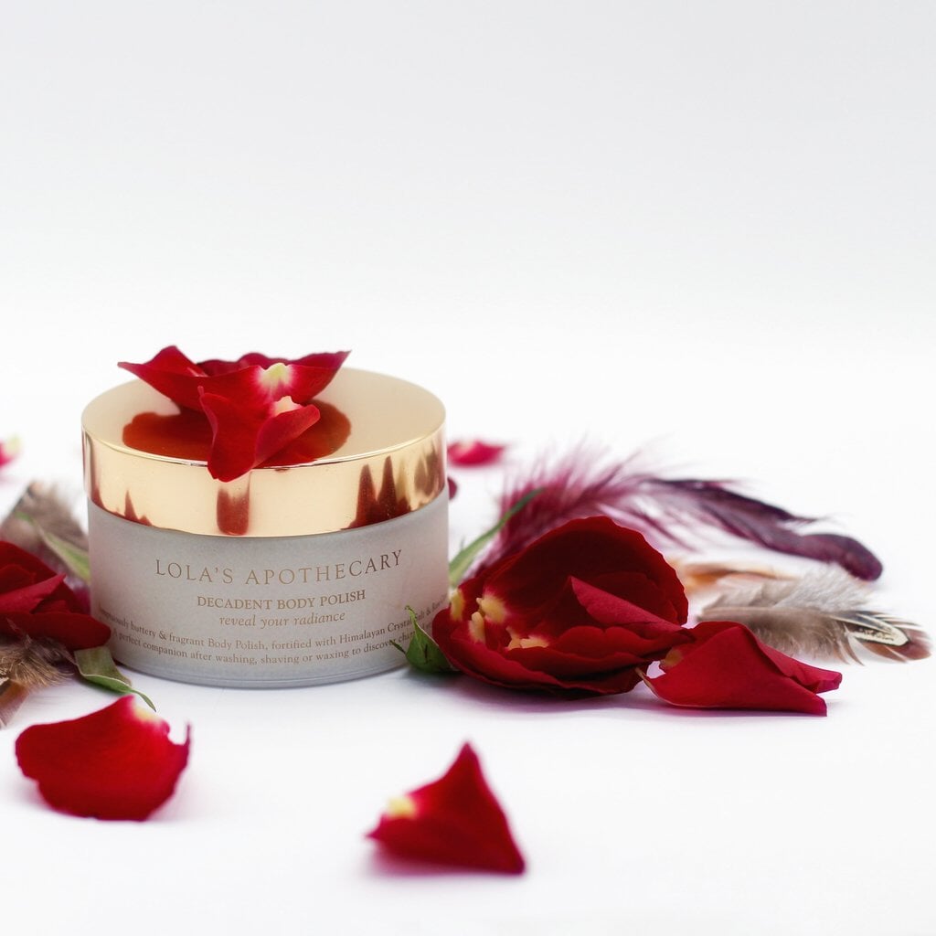 Glastiegel Lola's Apothecary Body Polish mit goldenem Deckel steht mit Rosenblättern und Federn dekoriert auf hellem Untergrund. North Glow