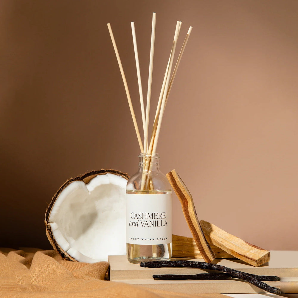 Sweet Water Decor Diffuser bottle with reed sticks in front of half a coconut with vanilla pods and sandalwood.