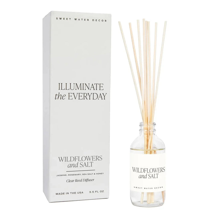 Transparente Flasche Diffuser Wildflowers and Salt von Sweet Water Decor mit Schilfrohrstangen, steht neben der Verpackung vor transparentem Hintergrund.