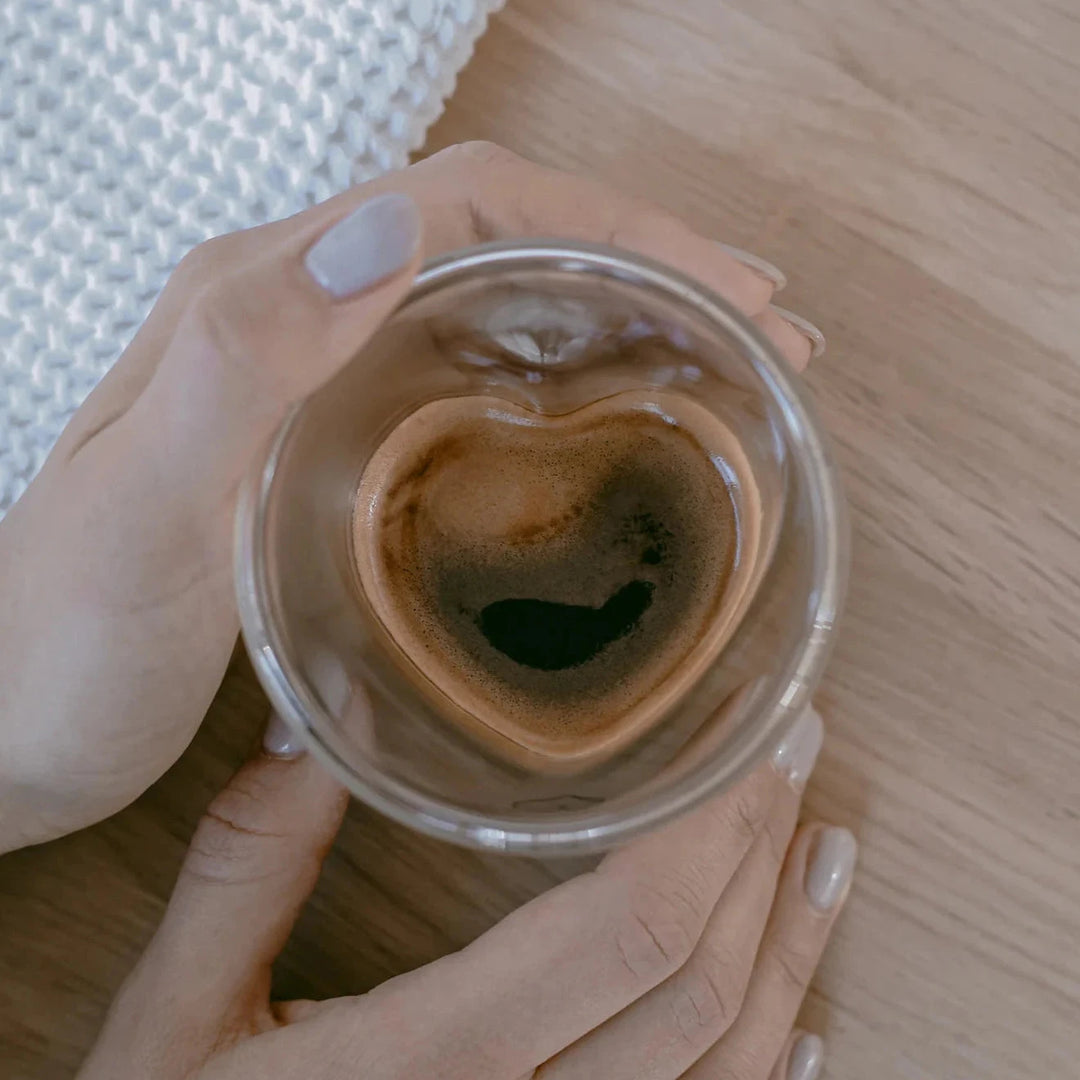 Heart-shaped glass with coffee is held by two hands, standing on a wooden table.