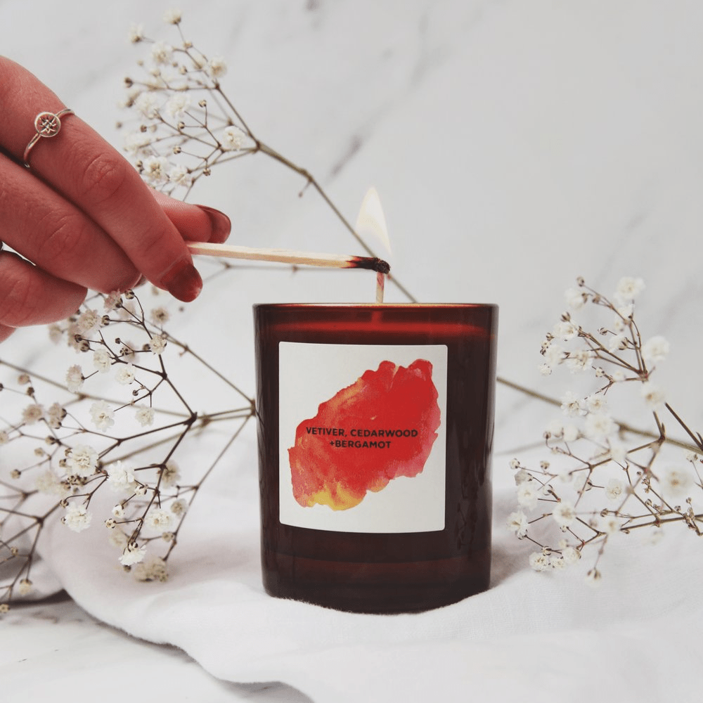 Self Care soy wax candle Cedarwood and Bergamot in brown glass, with a woman's hand lighting the candle
