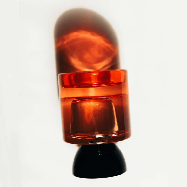 Vyrao Georgette glass bottle with black lid and reddish bottle lies upside down on a white background with a colored shadow in the background.