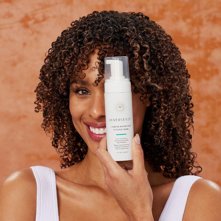 INNERSENSE I Create Definition Pump bottle held by a woman's hand in front of a woman's head with very curly hair.