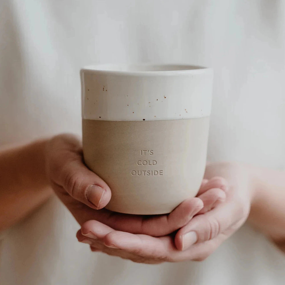 Coffee mug with embossed writing "It's cold outside" is held on two hands