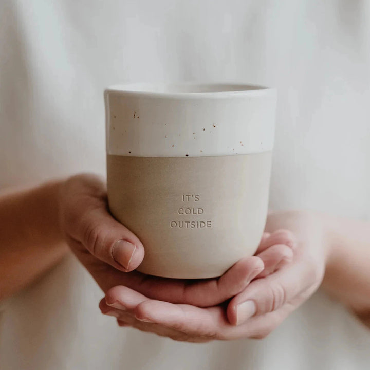 Coffee mug with embossed writing "It's cold outside" is held on two hands