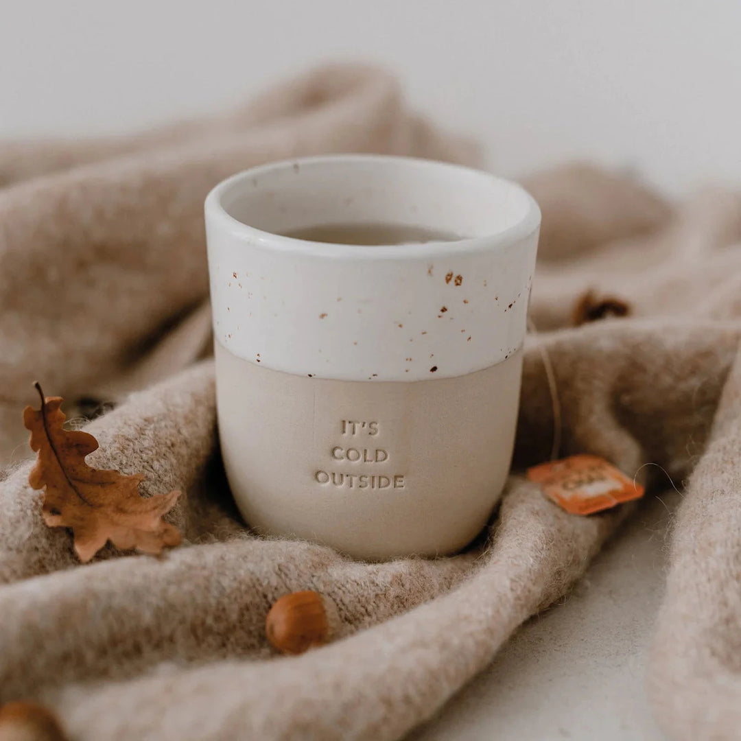 Coffee mug embossed with "it's cold outside" on a light brown blanket surrounded by an oak leaf and an acorn.