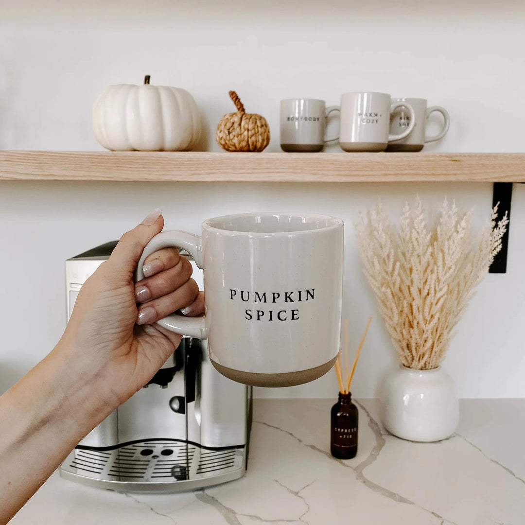Cremefarbene Kaffeetasse Pumpkin Spice von Sweet Water Decor wird von einer Hand vor einer Kaffeemaschine und einem Regal mit verschiedenen Tassen gehalten.