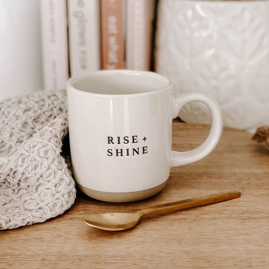 Cream colored coffee or tea cup with inscription Rise and Shine by Sweet Water Decor on a wooden table with decorative elements and spoon.