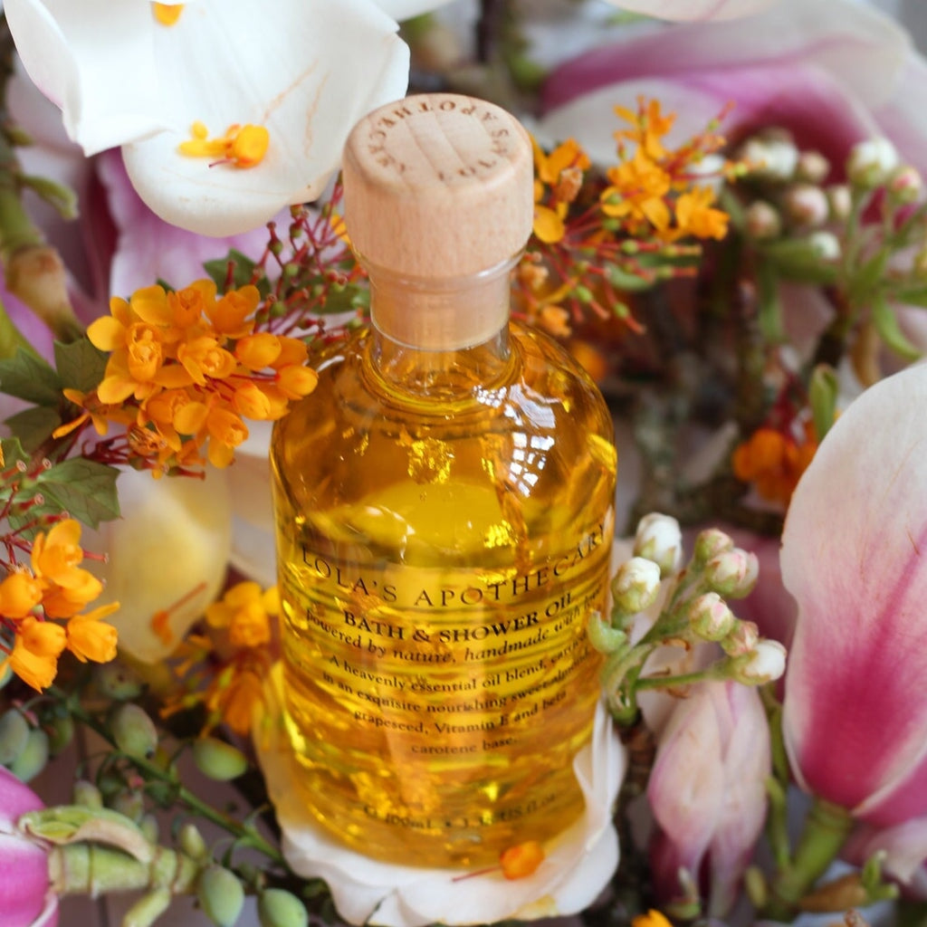 Eine Glasflasche Bath & Shower Oil von Lola's steht zwischen verschieden Büten. North Glow