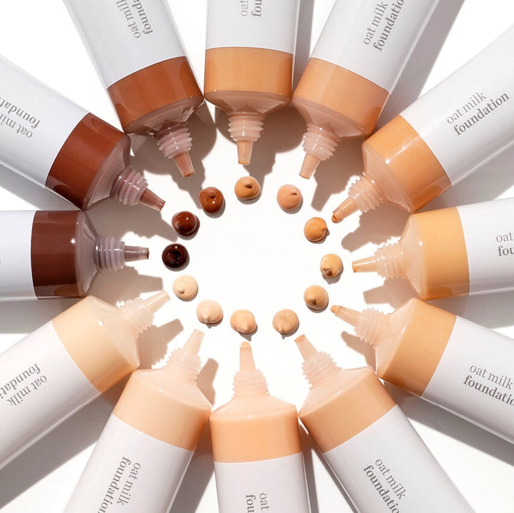 Various colors and tubes of Ere Perez Oat Milk Foundation placed in a circle, each with a drop of color and texture sample in the middle.