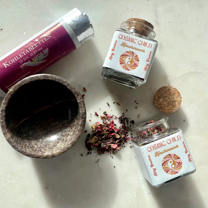 2 glass bottles of Cosmic Child incense with a cork stopper lie next to a soapstone bowl and a pack of charcoal tablets. 